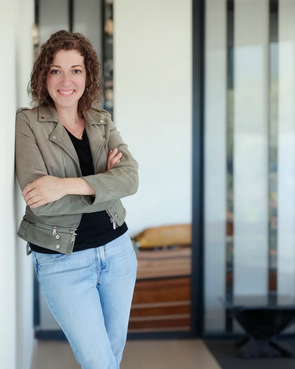 Céline, debout, adossée à un mur clair, bras croisés, veste kaki, sourit à l'objectif